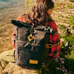 A JanSport Hatchet rolltop Adventure Backpack in black, worn by a teen on the edge of a riverbank.