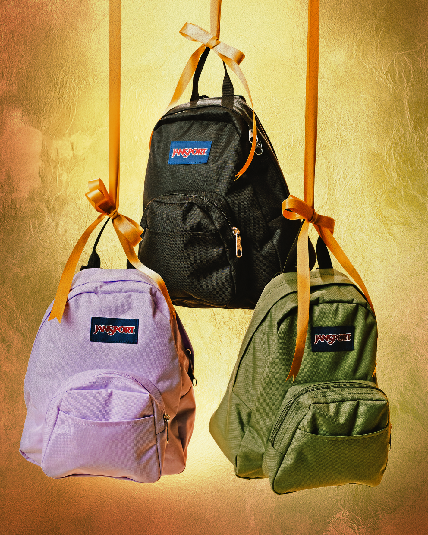 Three Jansport half pint backpacks in black, purple, and green with a visible brand logo on a textured gold background.