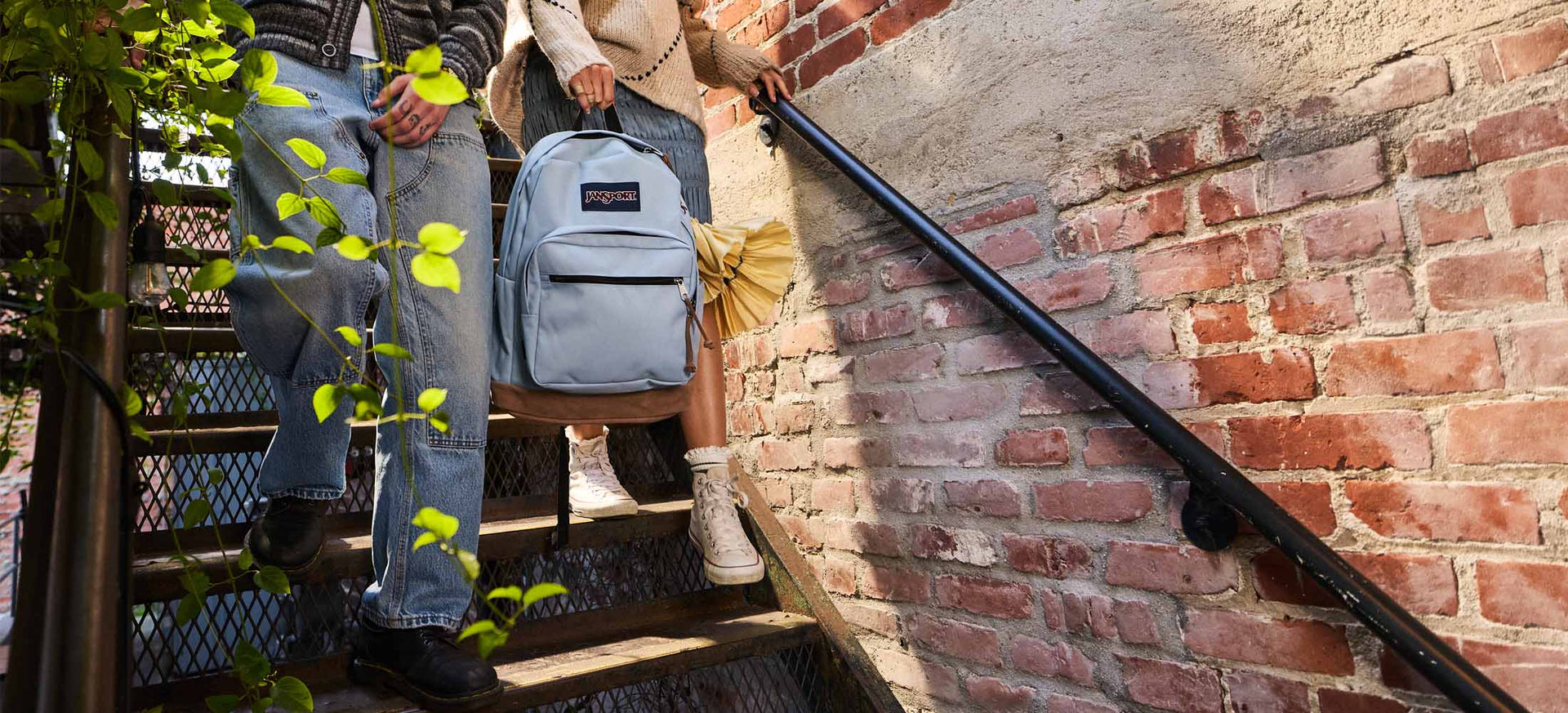 Two people walking up a set of stairs holding a Jansport rightpack with a brick wall in the background